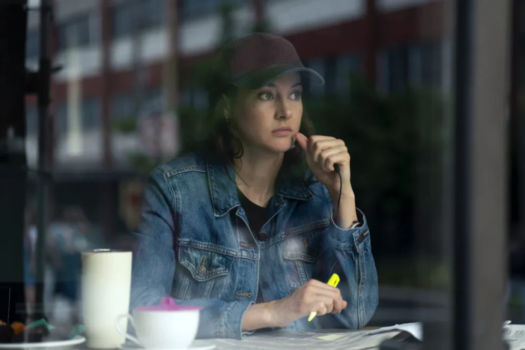 "Woman in denim jacket and baseball cap sits at a table inside a cafe, looking thoughtful while holding a yellow highlighter, with a mug and papers in front of her, city street visible through the window."