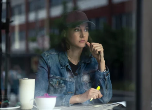 "Woman in denim jacket and baseball cap sits at a table inside a cafe, looking thoughtful while holding a yellow highlighter, with a mug and papers in front of her, city street visible through the window."